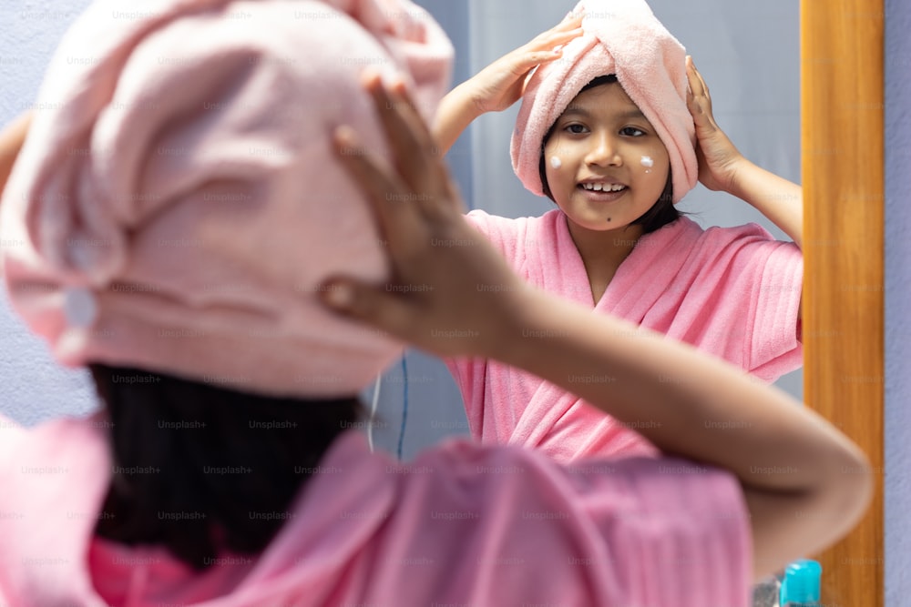 Ein süßes indisches Mädchen im rosa Bademantel trägt Gesichtscreme vor dem Spiegel auf