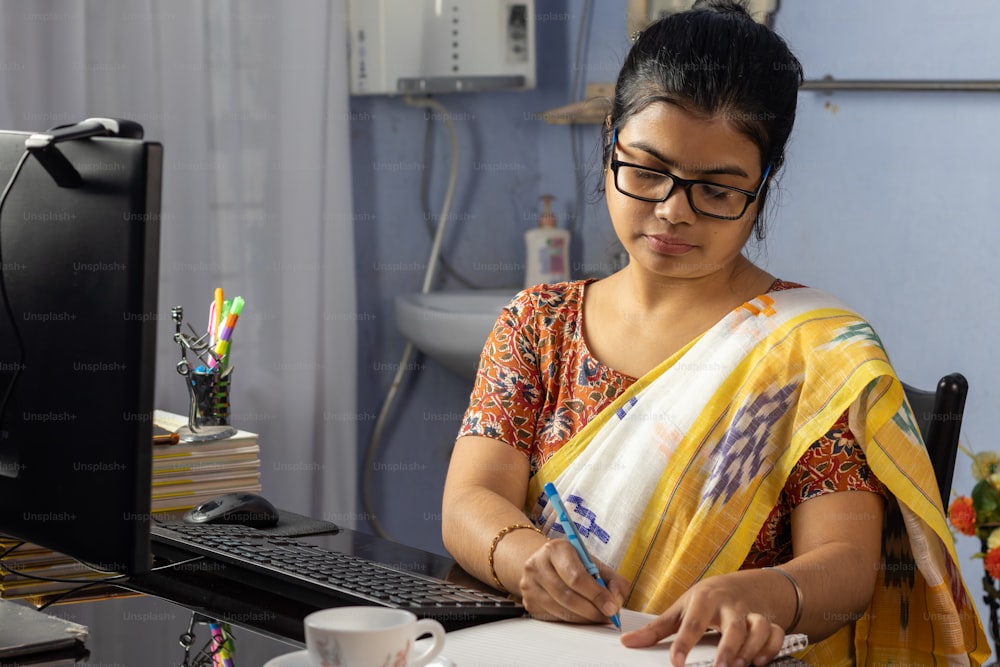사리를 입은 인도 여성이 컴퓨터로 작업하고 집에서 메모를 하고 집에서 일합니다.