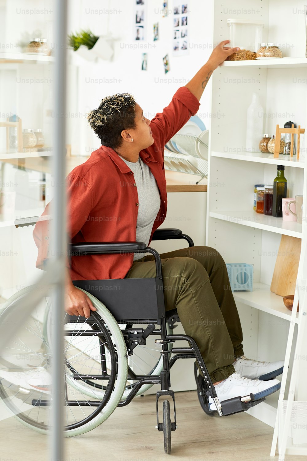 African woman with short hair sitting in wheelchair and trying to take product from the shelf at home
