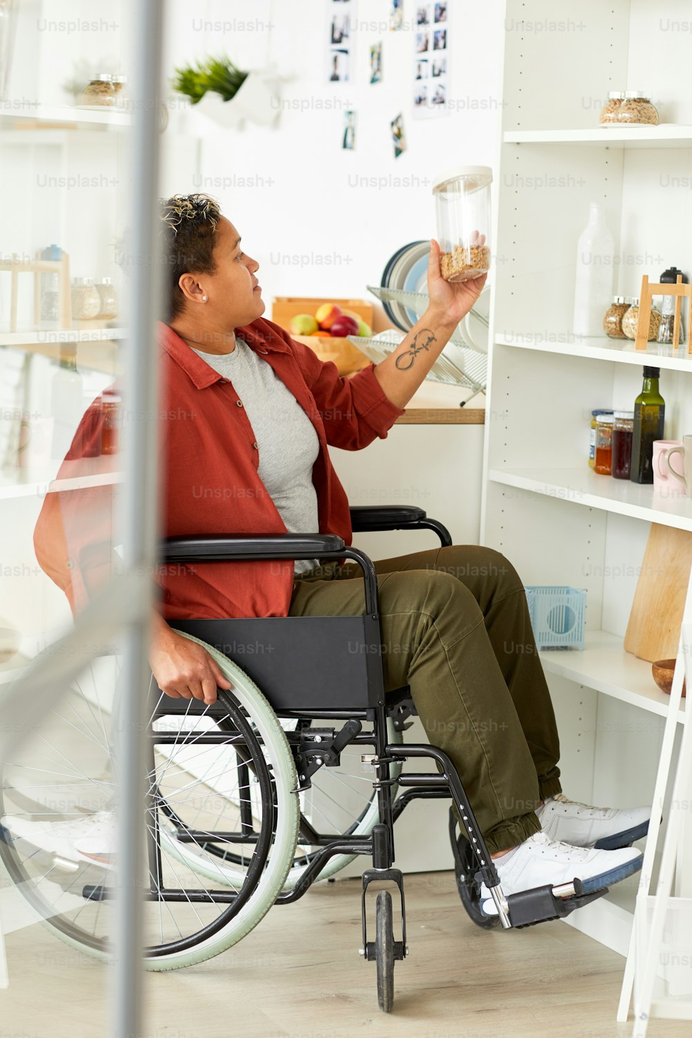 Mulher africana com deficiência sentada em cadeira de rodas perto das prateleiras e olhando para o produto em sua mão na cozinha