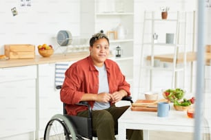 Retrato da mulher deficiente jovem africana sentada na cadeira de rodas e olhando para a câmera na cozinha em casa