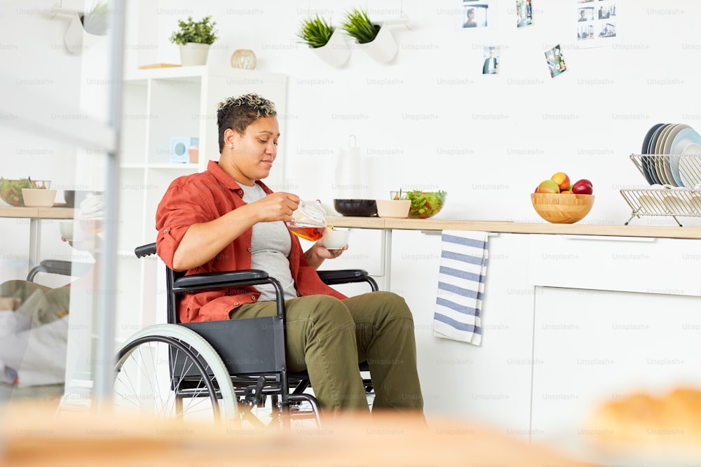 African disabled woman sitting in wheelchair and pouring tea in her cup from teapot in domestic kitchen
