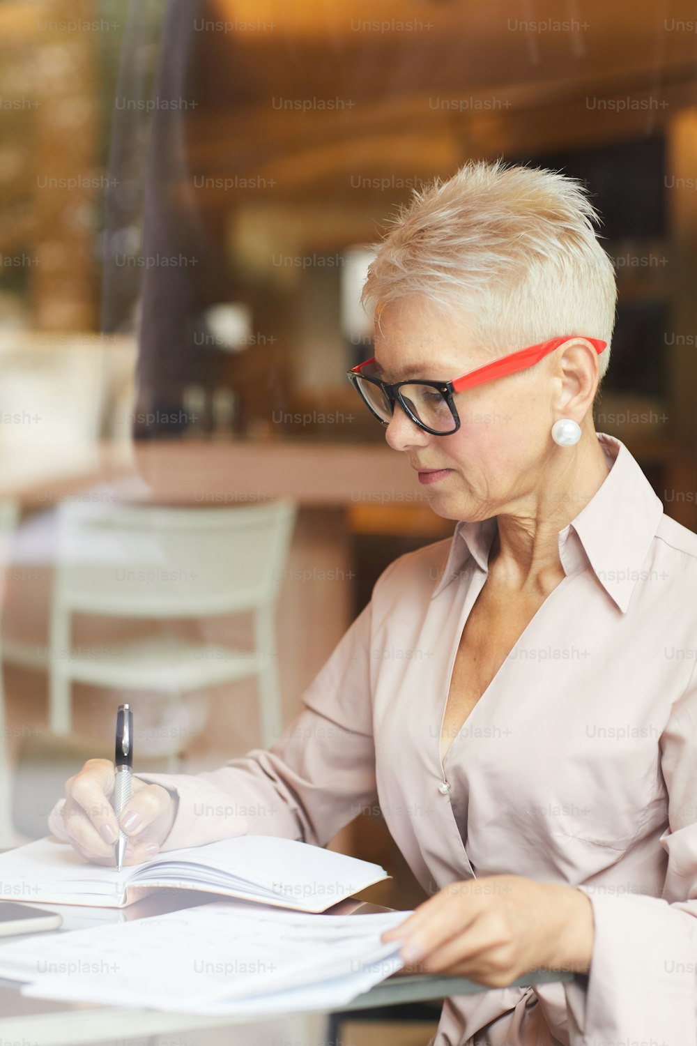 Donna d'affari matura concentrata con capelli corti biondi che indossa occhiali da vista seduta al tavolo e scrive qualcosa nei documenti nel caffè