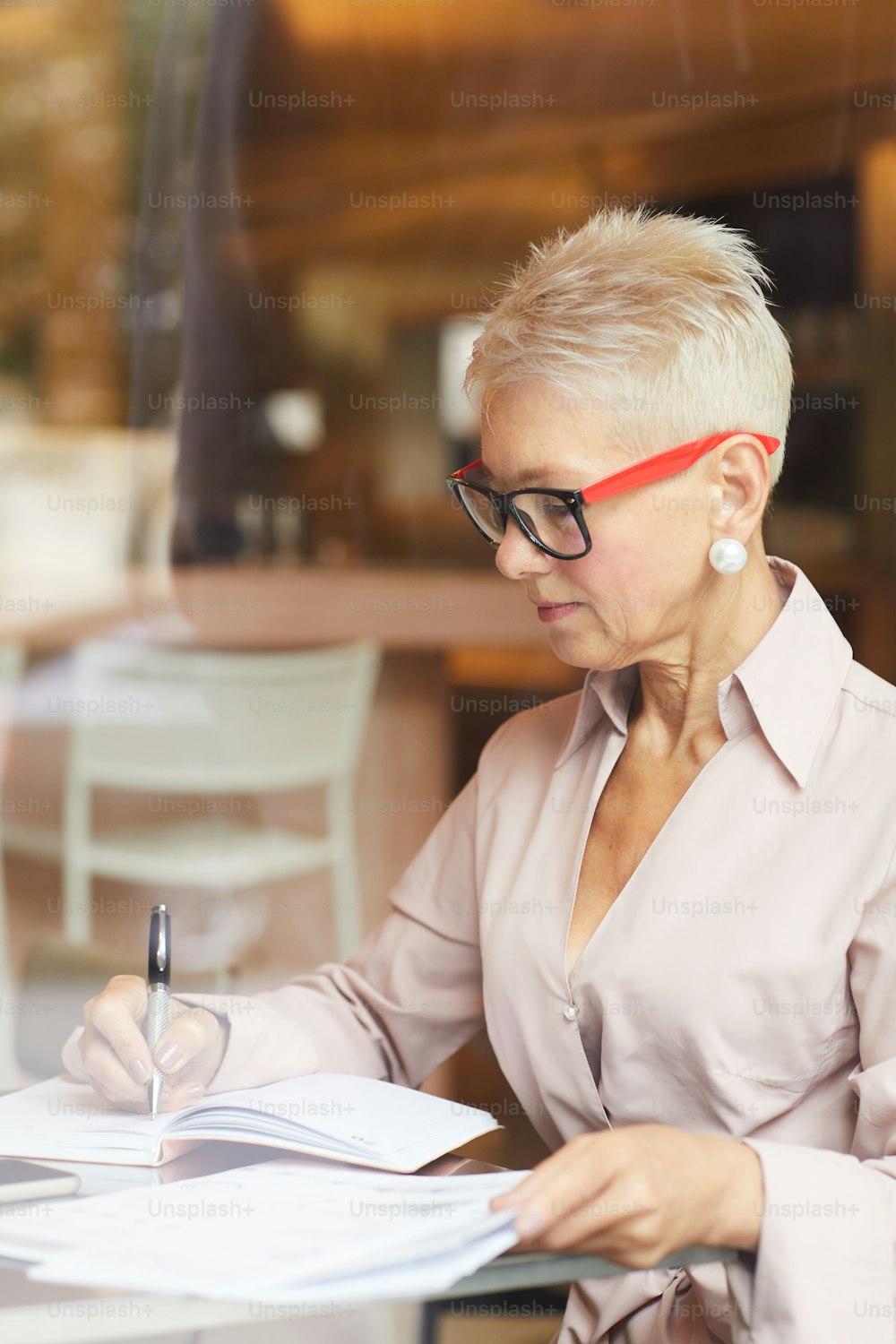 Concentrated mature businesswoman with blond short hair wearing eyeglasses sitting at the table and writing something in documents in cafe
