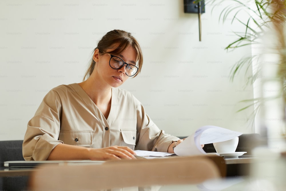 Giovane donna d'affari seria in occhiali che esamina i documenti mentre beve caffè nel caffè
