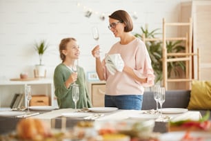 Young mother wiping the wineglasses with towel with her daughter helping her while they standing near the dining table at home