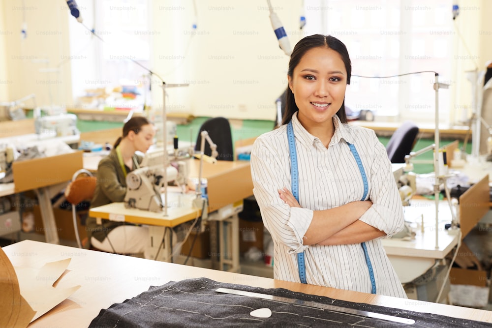 Portrait of Asian young woman smiling at camera while sewing clothing in workshop