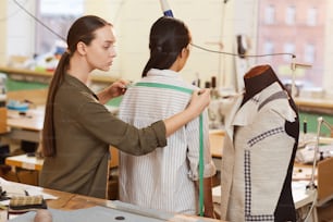 Young tailor measuring length of shoulders with tape measure of her customer while she standing back to her