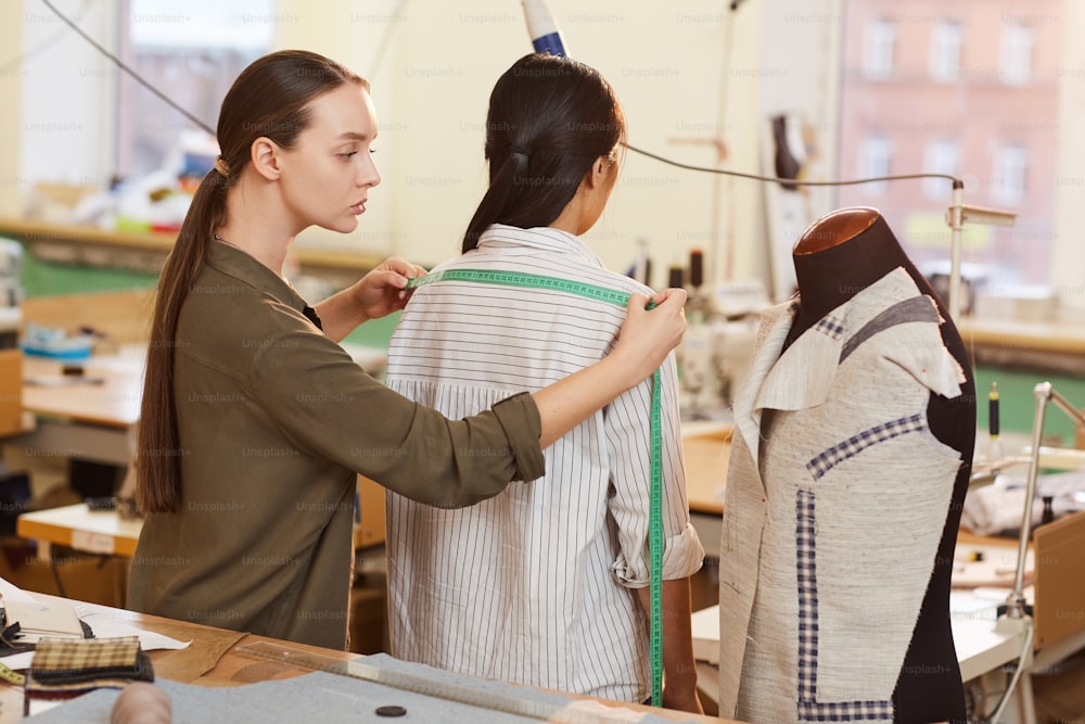 Jeune tailleur mesurant la longueur des épaules avec un ruban à mesurer de son client alors qu’elle se tient dos à elle