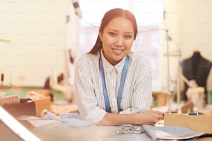Retrato de una sastre asiática sonriendo a la cámara mientras está parada cerca de su lugar de trabajo en el taller