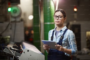 Portrait of female worker in eyeglasses and in overalls looking at camera while standing in the warehouse and using tablet pc