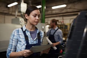 Young manual worker standing in overalls working online on tablet pc and looking at machine with her colleague working in the background