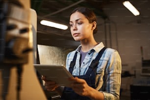Trabajadora en mono usando una tableta digital en su trabajo y observando el proceso mientras trabaja en la planta