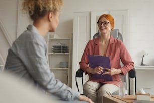 眼鏡をかけた赤い髪の女性が椅子に座って、オフィスでの心理療法中にクライアントに微笑んで話しかける