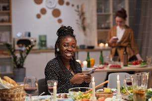 젊은 아프리카 여성이 휴대폰으로 테이블에 앉아 휴일 저녁을 먹으면서 카메라에 미소를 짓고 있다