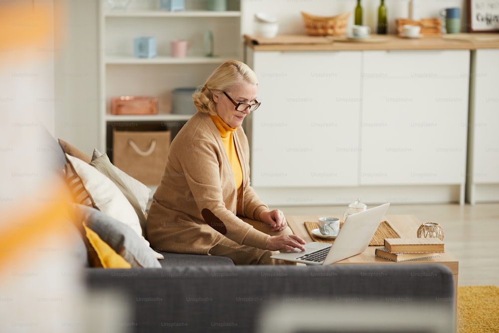 Mulher madura em óculos sentada no sofá e usando o computador portátil em seu trabalho on-line em casa
