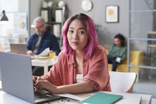 Retrato da mulher jovem asiática sentada na mesa e digitando no laptop no escritório
