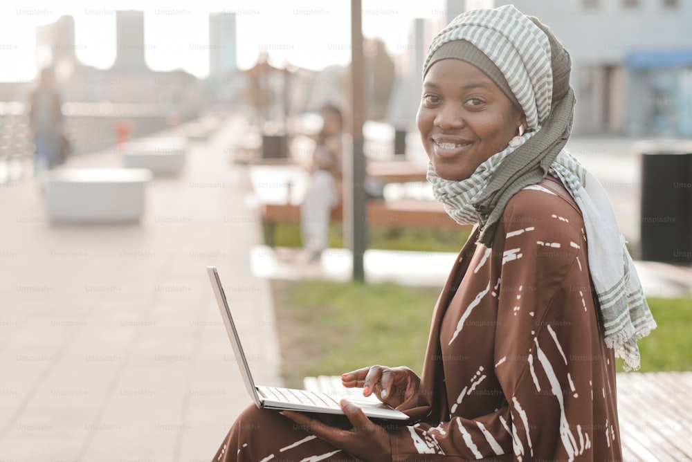 Portrait of African muslim woman smiling at camera while sitting on the bench and using laptop outdoors
