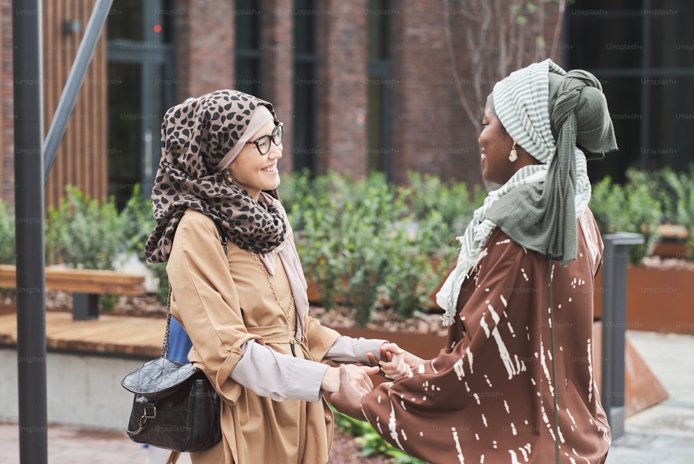 Two muslim women holding hands and smiling to each other during their meeting in the city