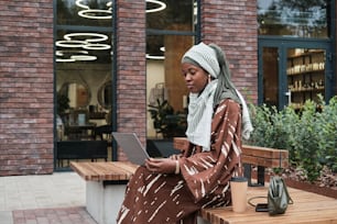 African muslim businesswoman sitting on the bench with laptop and workikng online outdoors