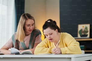 Young Caucasian teacher sitting at table next to female student with Down syndrome helping with subject during individual class