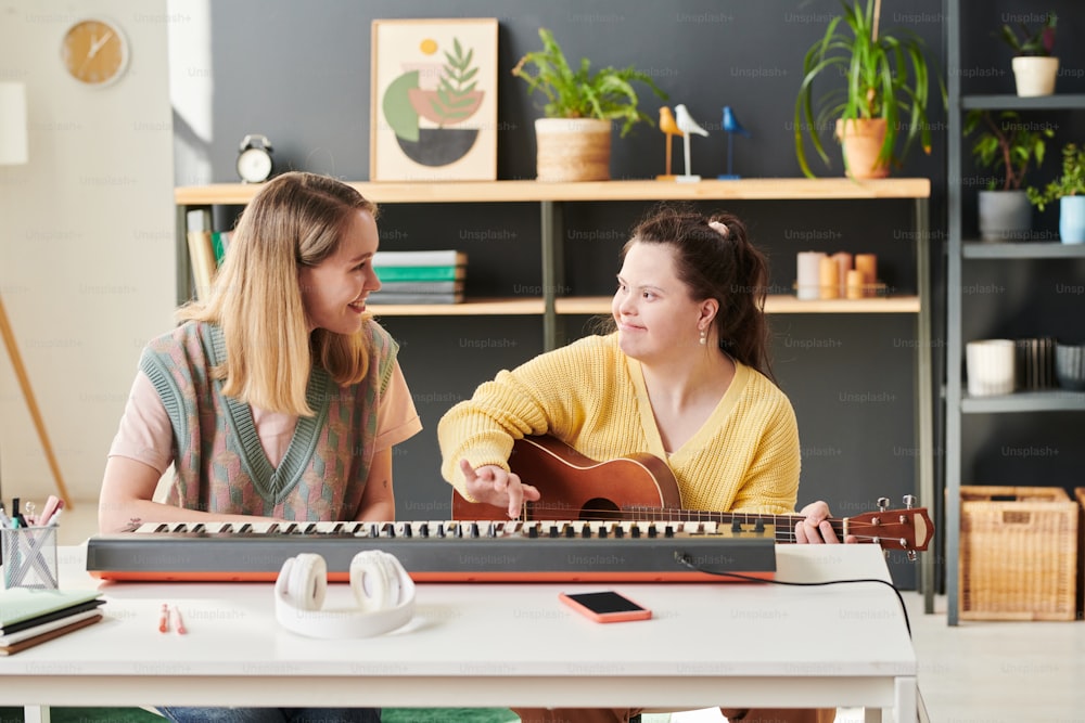 Jeune femme de race blanche assise à côté de son élève atteinte du syndrome de Down lui apprenant à jouer de la guitare