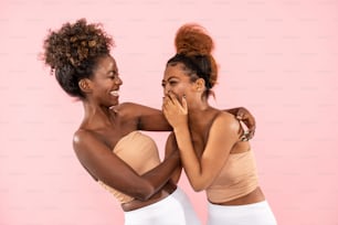 Studio shot of happy Afro female sisters enjoying good moment, chatting and having fun together during free time, isolated over pink pastel studio background. Friendship and real people emotions concept.