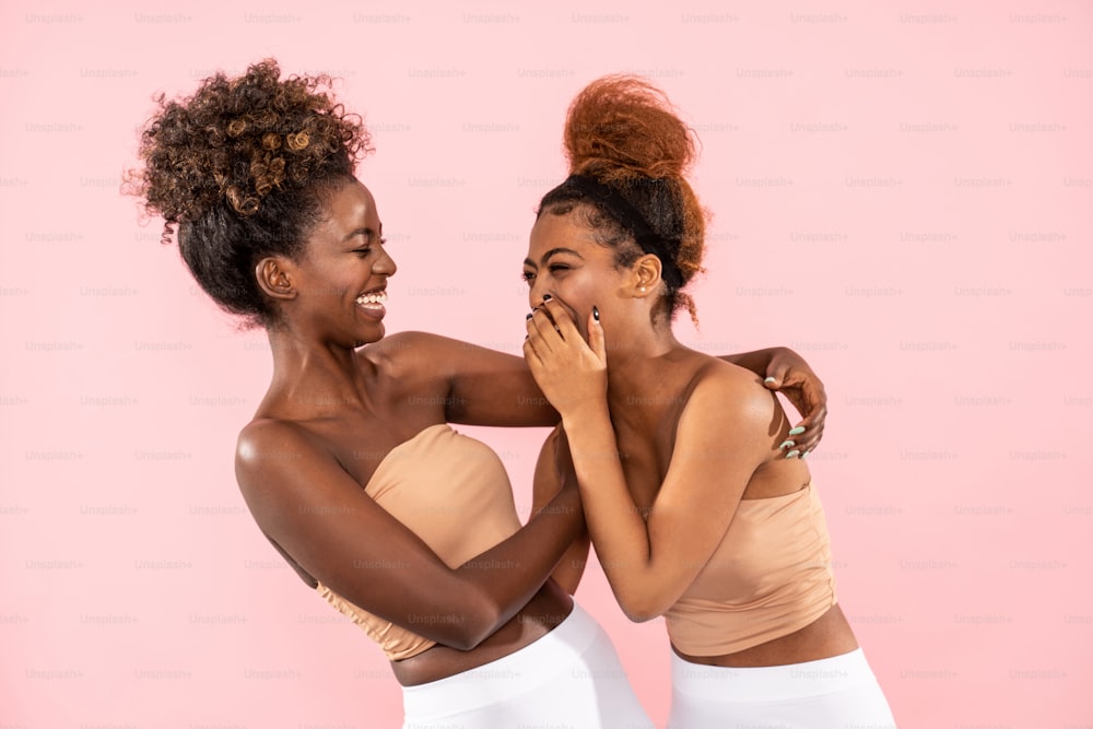 ピンクのパステルスタジオ背景に、幸せなアフロの女性姉妹が楽しい瞬間を楽しんだり、自由時間に一緒におしゃべりしたり楽しんだりするスタジオショット。友情と実在の人々の感情のコンセプト。