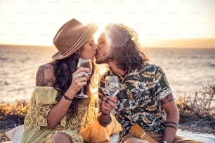 Fashionable boho couple with wine having romantic date on sunset beach, kissing, spending time together. Real people emotions and love concept.