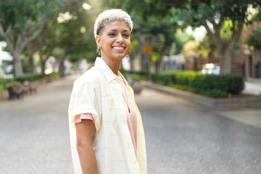 Portrait of natural millenial woman with short blonde hairstyle wearing silver jewelry, earrings and necklace, smiling and looking to the camera. Beautiful girl walking on sunny summer day. Real people emotions. Lifestyle.