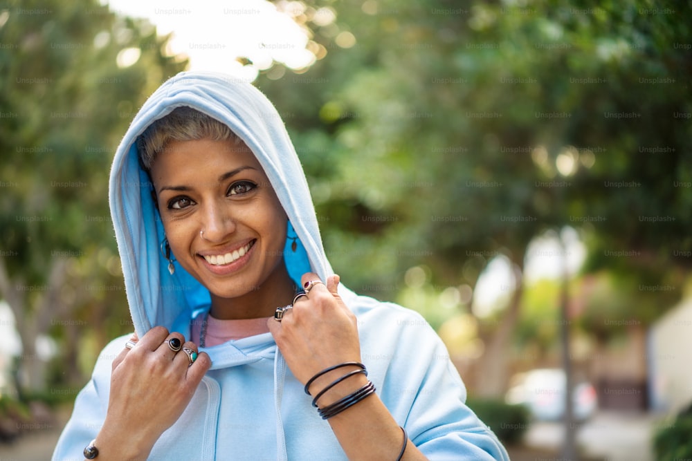 Heureuse jeune femme hispanique souriante aux yeux bruns regardant la caméra. Fille portant un sweat à capuche bleu à la mode et beaucoup de bagues. Photo extérieure dans le parc.