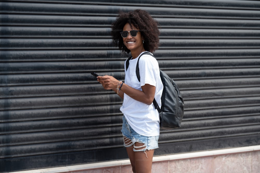 Chica atractiva y feliz en la calle de la ciudad usando el teléfono móvil, revisando las redes sociales y los correos electrónicos. Mujer con peinado afro con ropa elegante y de moda. Emociones de personas reales.