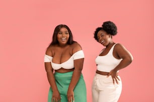 Studio shot of happy dark skinned, young sisters enjoying good moment, dancing and having fun together, posing over pink pastel studio background. Friendship and real people emotions concept. Body positive. Copy space.