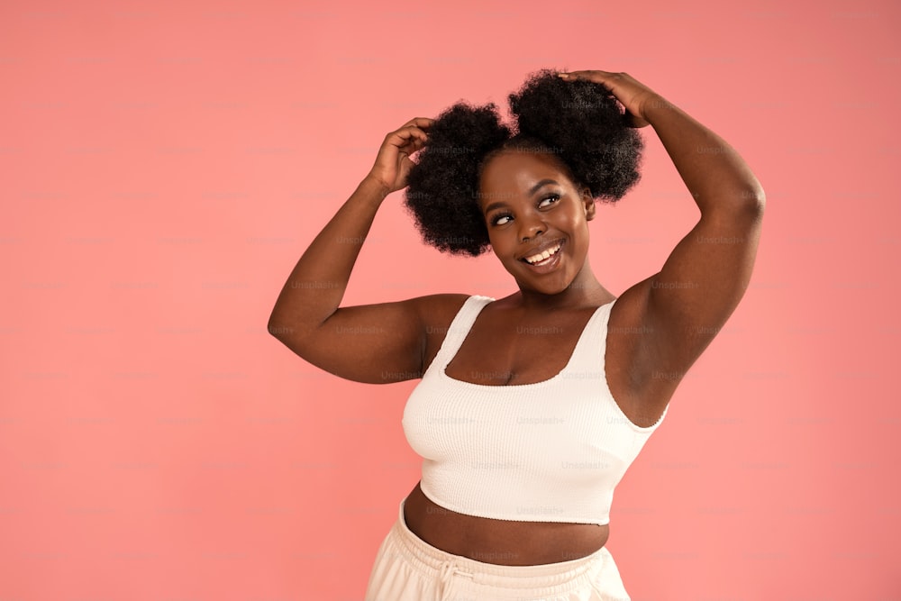 Cute Plus Size african Woman with natural makeup and fashionable hairstyle posing on a pastel pink Studio Background. Real people emotions, toothy smile. Young girl in white crop top. Body positive. A lot of copy space.