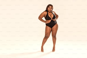 Full-length photo of beautiful plus size dark skinned woman posing on light gray, wearing black fashionable swimsuit. Body positive, conscious concept.