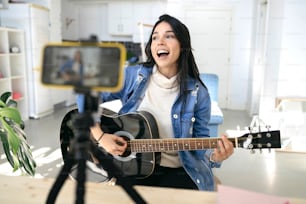 Giovane donna influente che suona la chitarra durante il podcast o la trasmissione video in diretta per il pubblico dal cellulare di casa - Concept Art, hobby e video blog