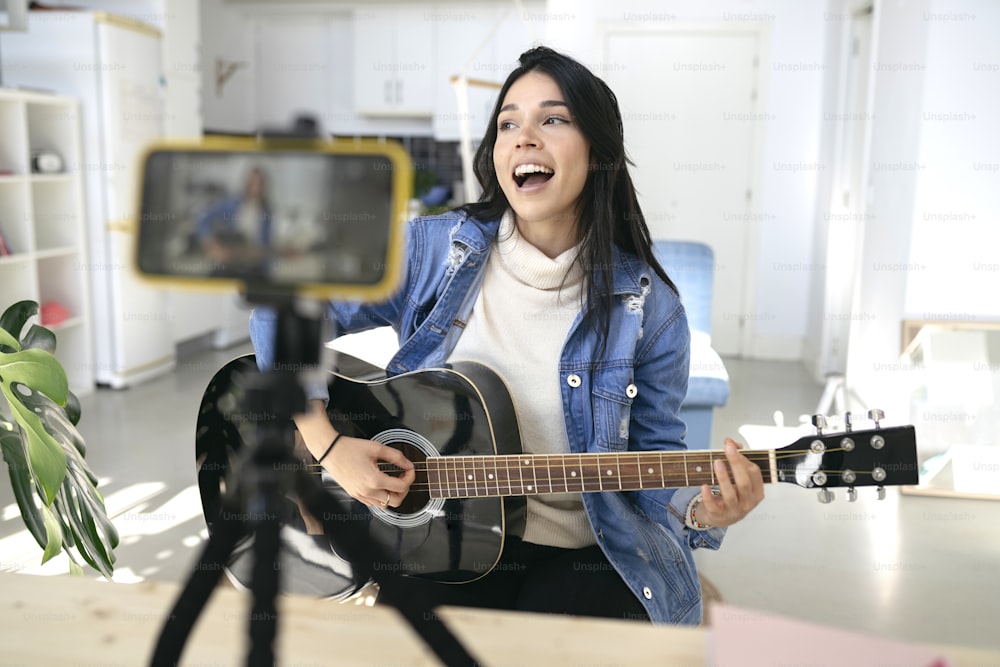 Jeune femme influente jouant de la guitare lors d’un podcast ou d’une diffusion vidéo en direct pour le public depuis le téléphone portable à la maison - concept art, hobby et blog vidéo