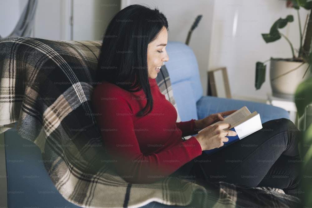 Hermosa mujer hispana leyendo un libro sentada en el sofá de su casa relajándose con una manta - concepto de entretenimiento en el hogar -