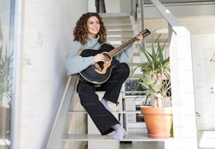 lockige junge Frau, auf der Treppe des Hauses, spielt Gitarre und blickt auf die Sonne