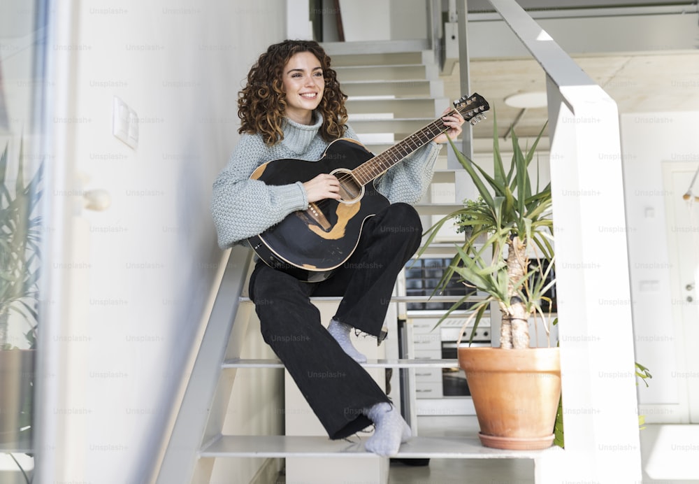 lockige junge Frau, auf der Treppe des Hauses, spielt Gitarre und blickt auf die Sonne