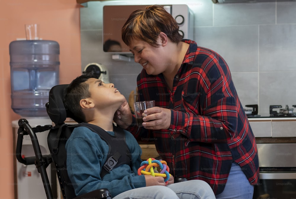 Hispanic mother caring for wheelchair-bound disabled son in the kitchen, giving him a drink while smiling.