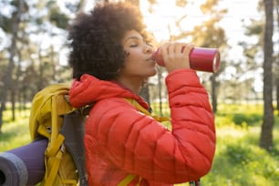 Mujer afroamericana tomando un descanso para beber de una botella de agua mientras camina por la naturaleza al atardecer - Concepto de senderismo deportivo -