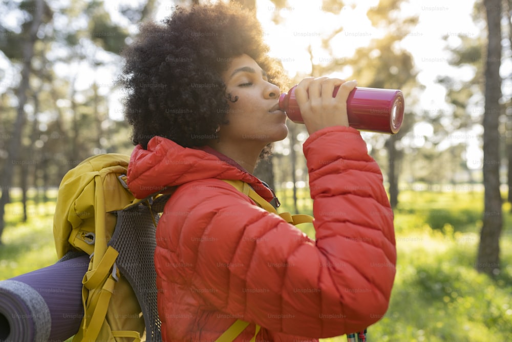 Donna afroamericana che fa una pausa per bere da una bottiglia d'acqua durante un'escursione nella natura al tramonto - Concetto di escursionismo sportivo -