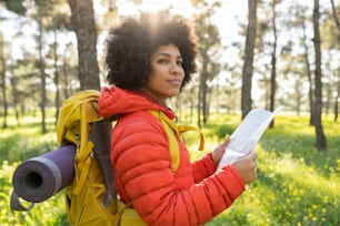 Caminhante atraente mulher afro-americana com mochila de caminhada na floresta ao pôr do sol, segurando um mapa - conceito de trekking -