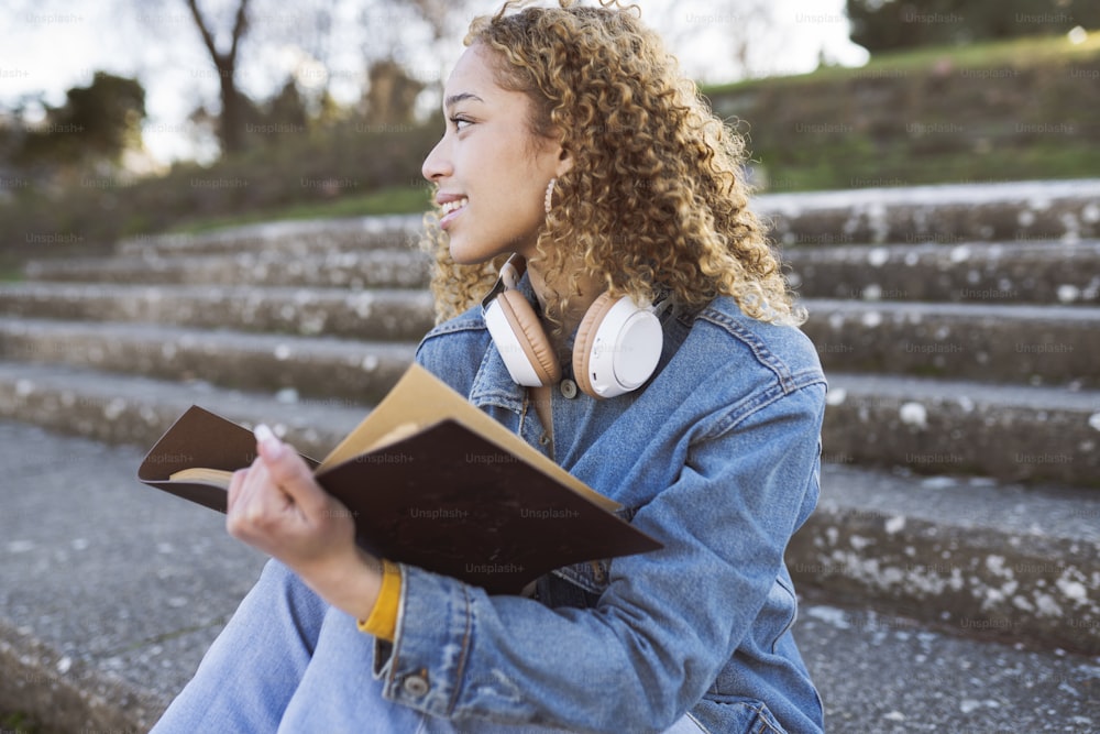 curly-haired hispanic woman, reading a book on park stairs, with denim clothes and headphones on her neck