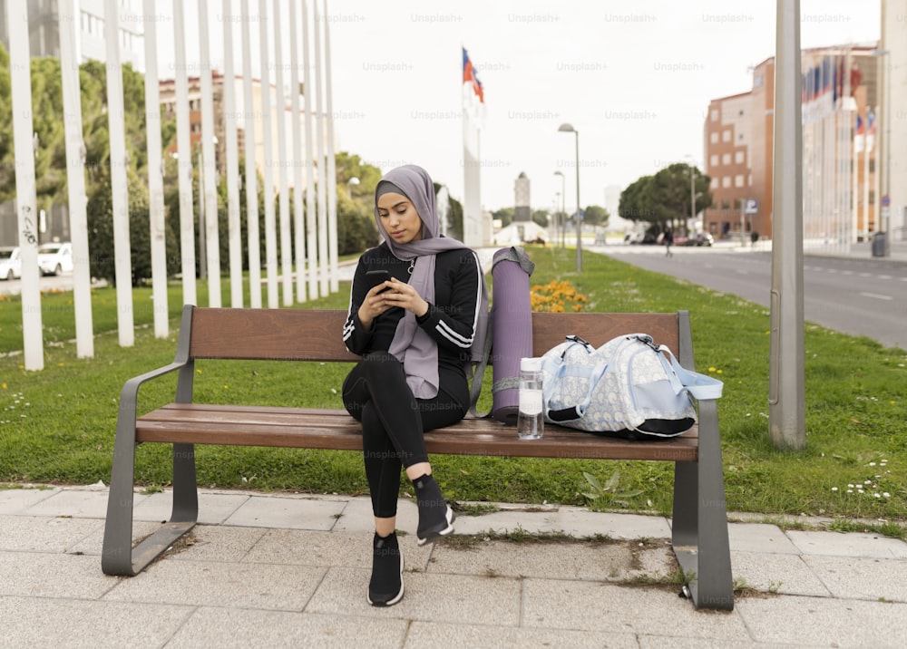 islamic woman sitting on a park bench with smartphone, she is wearing sportswear.