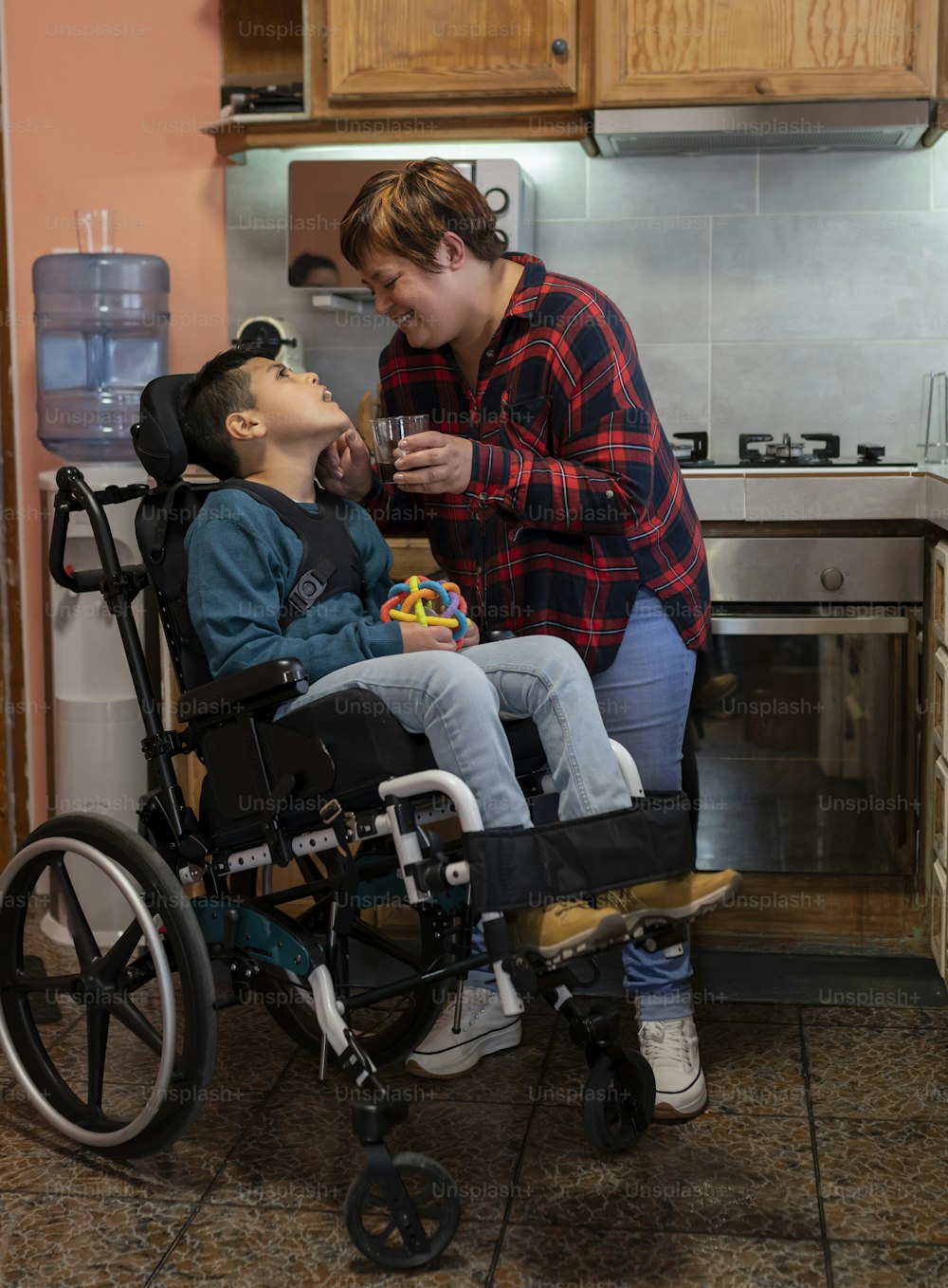 Madre che si prende cura del figlio disabile costretto su una sedia a rotelle in cucina. - concetto di assistenza sanitaria e disabilità -