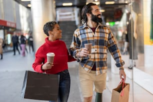 casal multirracial fazendo compras na cidade olhando para a vitrine, enquanto carrega sacolas e café para ir, dia dos namorados