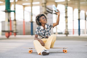 スケートボードと眼鏡をかけたきれいな金髪のアフロ女性、座ってスマートフォンで写真を撮る