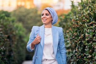 Front view of a smiling muslim woman standing among large bushes wearing a blue light suit and headscarf holding her purse on a pathway looking at camera in a sunny day.